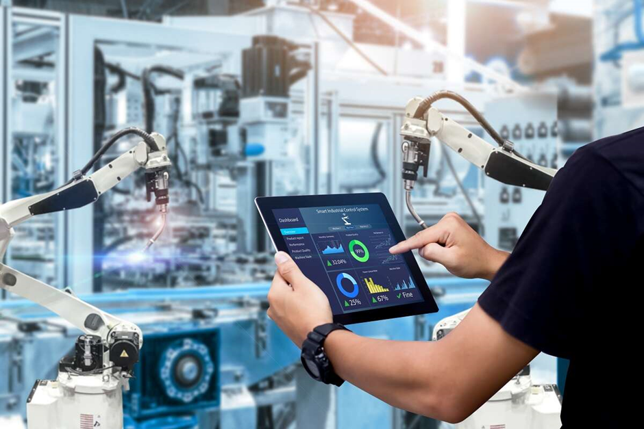 A person uses a tablet to monitor robotic arms in a factory, with the tablet displaying a dashboard of smart industrial system metrics, including charts and performance indicators.